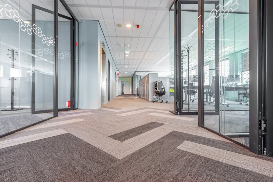 Interior view of a modern office space in the Bentall Centre Kingston with glass partition walls and tiles. The corridor has a patterned carpet in shades of gray and beige, appearing clean and well-maintained. Office chairs and desks are visible through the glass walls, with natural light illuminating the area, reflecting a professional environment. The image showcases surface cleaning and maintenance of flooring and glass surfaces, representative of professional cleaning services by Kingstonuponthamescarpetcleaners.co.uk. The space appears tidy, organized, and hygienic, emphasizing deep cleaning and surface sanitisation for commercial premises.