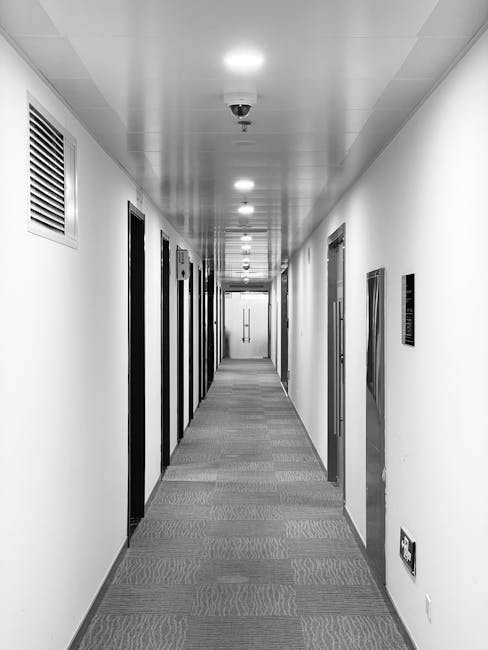 A long, straight corridor in a commercial building featuring smooth, white-painted walls and a textured grey carpeted floor. The ceiling is fitted with evenly spaced recessed lighting fixtures providing bright illumination, while a security camera is mounted centrally on the ceiling. On the left side, there are several closed doors with black frames, and a ventilation grille is visible near the top of the wall. On the right side, additional doors and an electrical outlet near the floor are present. The surface appears clean and well-maintained, reflecting professional deep cleaning standards typical of office environments, as recommended by Kingstonuponthamescarpetcleaners.co.uk for hygienic surface cleaning and maintenance.