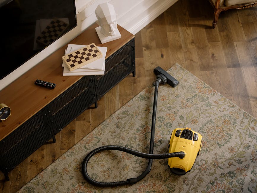 Interior living room scene featuring a hardwood floor with a light-colored patterned rug, vacuum cleaner with a yellow body and black hose positioned on the rug near a black metal media console, which holds a flat-screen TV, a white bust sculpture, a chessboard, a few books or papers, and a remote control. The room is well-lit, with a white baseboard along the wall, and a brown-colored wooden armchair partially visible in the top right corner, suggesting a clean and maintained space suitable for surface cleaning and sanitisation as part of domestic cleaning services from kingstonuponthamescarpetcleaners.co.uk.