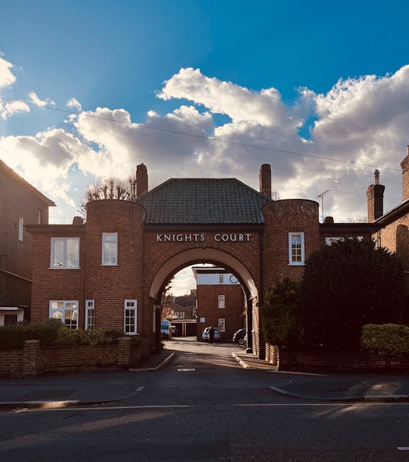 A brick archway labeled 'KNIGHTS COURT' forms the entrance to a residential area in Kingston upon Thames, featuring a dark tiled roof and two chimney stacks. The archway is flanked by two symmetrical brick buildings with white-framed windows, and lush green bushes border the site. The scene is illuminated by natural daylight, with a partly cloudy sky overhead, showcasing fluffy white clouds and patches of blue sky. This image exemplifies the exterior of a well-maintained residential development, illustrating structures likely subjected to surface cleaning and maintenance by Kingstonuponthamescarpetcleaners.co.uk, emphasizing the importance of thorough exterior and surface cleaning for enhancing property appearance and hygiene.