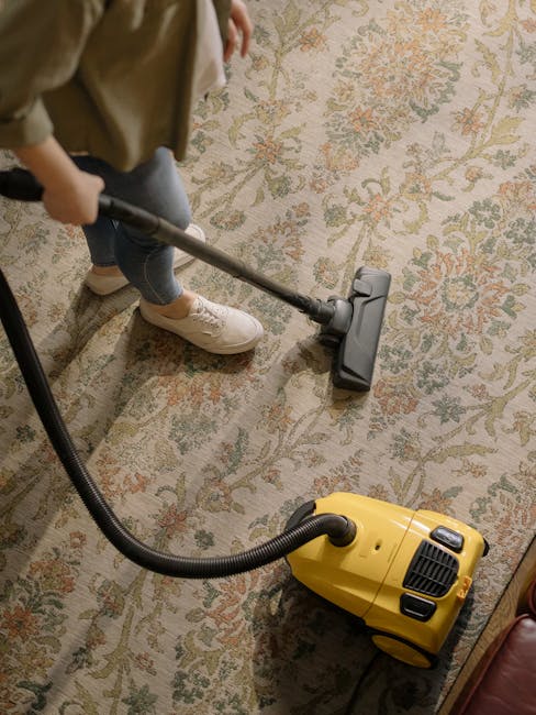 Interior living room scene featuring a hardwood floor with a light-colored patterned rug, vacuum cleaner with a yellow body and black hose positioned on the rug near a black metal media console, which holds a flat-screen TV, a white bust sculpture, a chessboard, a few books or papers, and a remote control. The room is well-lit, with a white baseboard along the wall, and a brown-colored wooden armchair partially visible in the top right corner, suggesting a clean and maintained space suitable for surface cleaning and sanitisation as part of domestic cleaning services from kingstonuponthamescarpetcleaners.co.uk.